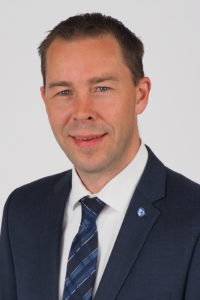 Professionelles Porträt eines Mannes in Anzug mit Krawatte, lächelnd vor neutralem Hintergrund. Ideal für Unternehmensprofile oder persönliche Branding.