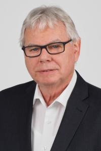 Porträt eines älteren Mannes mit Brille, der einen schwarzen Blazer und ein weißes Hemd trägt, vor einem neutralen Hintergrund.