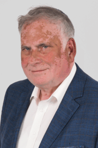 Ein Mann mit weißem Haar und blauem Hemd. Er lächelt.