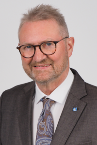 Professionelles Porträt eines Mannes mit Brille, Anzug und Krawatte, lächelnd vor neutralem Hintergrund.