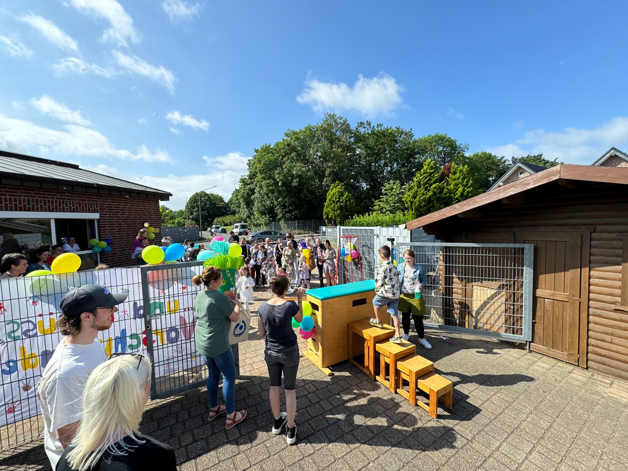 Menschenmenge bei einem Fest im Freien, umgeben von bunten Dekorationen und Ständen, mit Bäumen im Hintergrund und strahlendem Sonnenschein.