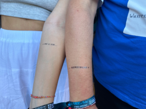 Zwei Arme mit minimalistischen Tattoos sind sichtbar, die jeweils unterschiedliche Schriftzüge auf den Unterarmen zeigen. Die Personen tragen lässige Kleidung, was einen entspannten, freundschaftlichen Moment vermittelt.