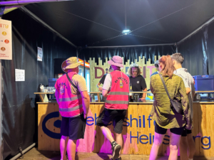 Besucher in einem Festivalzelt, die an einem Infostand mit freundlichem Personal stehen, während sie Informationen über das Event erhalten.