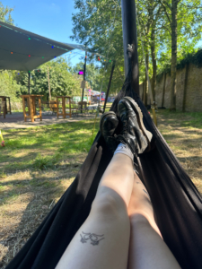 Person entspannt in einer Hängematte im Freien, umgeben von Bäumen und einer sonnigen Landschaft.