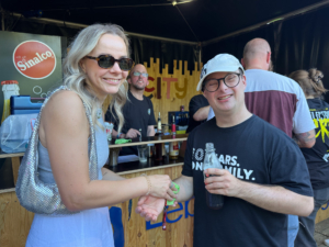 Zwei Personen stehen vor einer Bar. Eine trägt eine silberne Kette und die andere einen Hut.