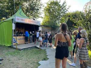 Menschen stehen in einer Schlange vor einem Verkaufsstand auf einem Festival im Freien, umgeben von Bäumen und sonnigem Wetter.