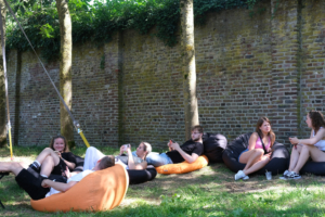 Mehrere Personen sitzen und liegen auf Luftmatratzen im Gras vor einer Mauer.