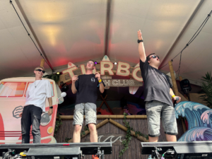 Zwei Männer stehen auf einer Bühne. Einer hebt die Arme. Hinter ihnen ist ein Banner mit einem Wellenmuster zu sehen.