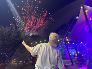 Feuerwerk am Abend. Ein Mann steht mit dem Rücken zum Betrachter und zeigt nach oben. Es gibt Musik und viele Lichter.