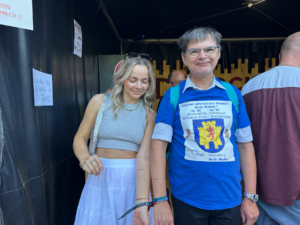 Zwei Personen stehen vor einem Veranstaltungsort, die Frau trägt ein graues Crop-Top und einen weißen Rock, der Mann trägt ein blaues T-Shirt mit einem Wappen. Sie lächeln und scheinen sich auf die Veranstaltung zu freuen.