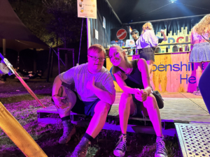 Junge und Mädchen sitzen freundlich zusammen auf einer Holzbank vor einem bunten Imbissstand bei Nacht, umgeben von Lichtern und festlicher Atmosphäre.