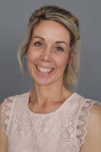Eine Frau lächelt. Sie hat blonde Haare und trägt ein weißes Oberteil.