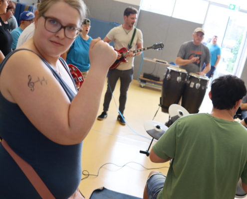 Eine Frau steht und spielt Gitarre. Hinter ihr sitzen Leute. Sie hören Musik.