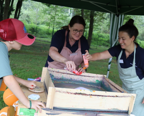 Zwei Personen arbeiten mit Farbe in einer Kiste. Eine Person hält die Kiste, die andere gibt Farbe dazu.