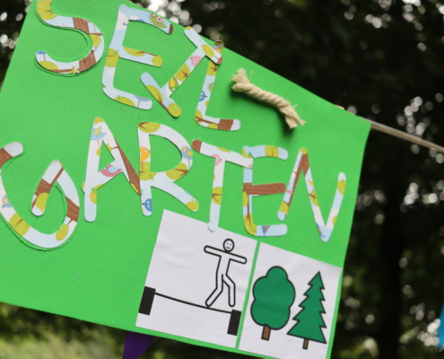 Buntes Schild mit der Aufschrift "Mein Garten", verziert mit bunten Materialien und Bildern von Bäumen und einer Figur, das in einem Garten hängt.