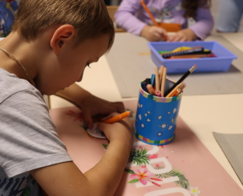 Ein Kind sitzt am Tisch und malt mit Buntstiften. Es gibt einen Stiftekübel und ein Tablett mit Farben auf dem Tisch.