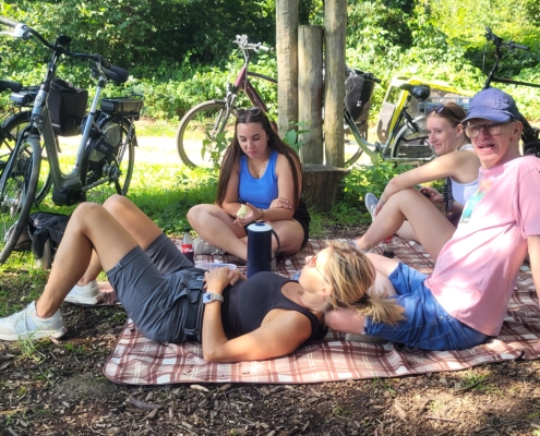 Gruppe von Freunden entspannt auf einer Picknickdecke im Freien, umgeben von Fahrrädern und Bäumen, während sie die Sonne genießen.