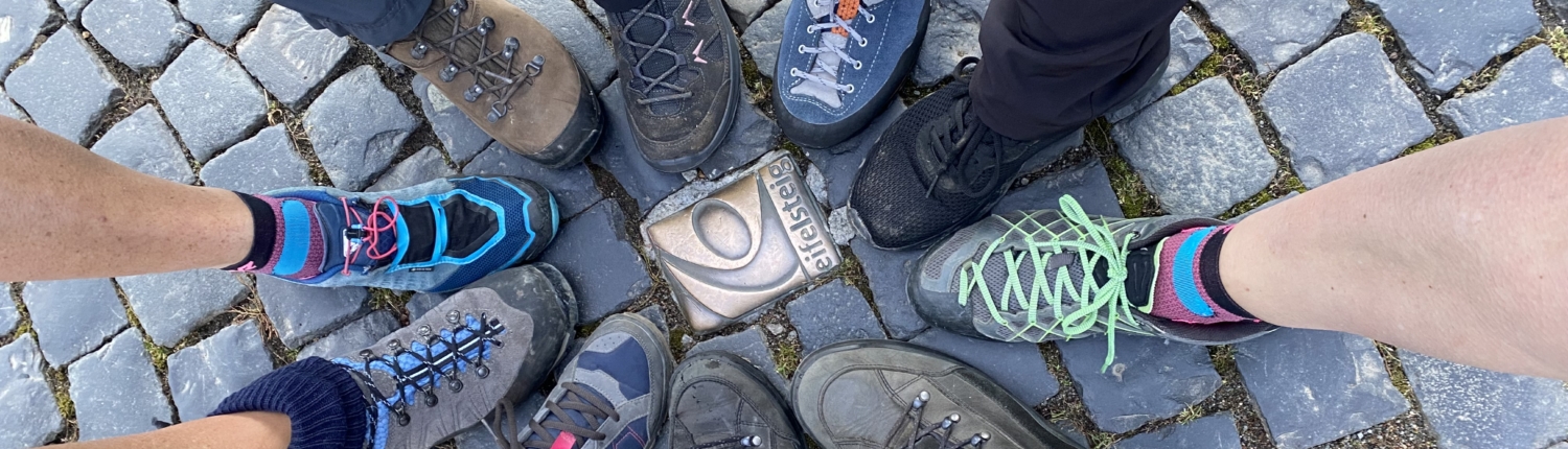 Verschiedene Wanderschuhe und Socken von mehreren Personen stehen auf einem gepflasterten Boden, was auf eine gemeinsame Outdoor-Aktivität hinweist.