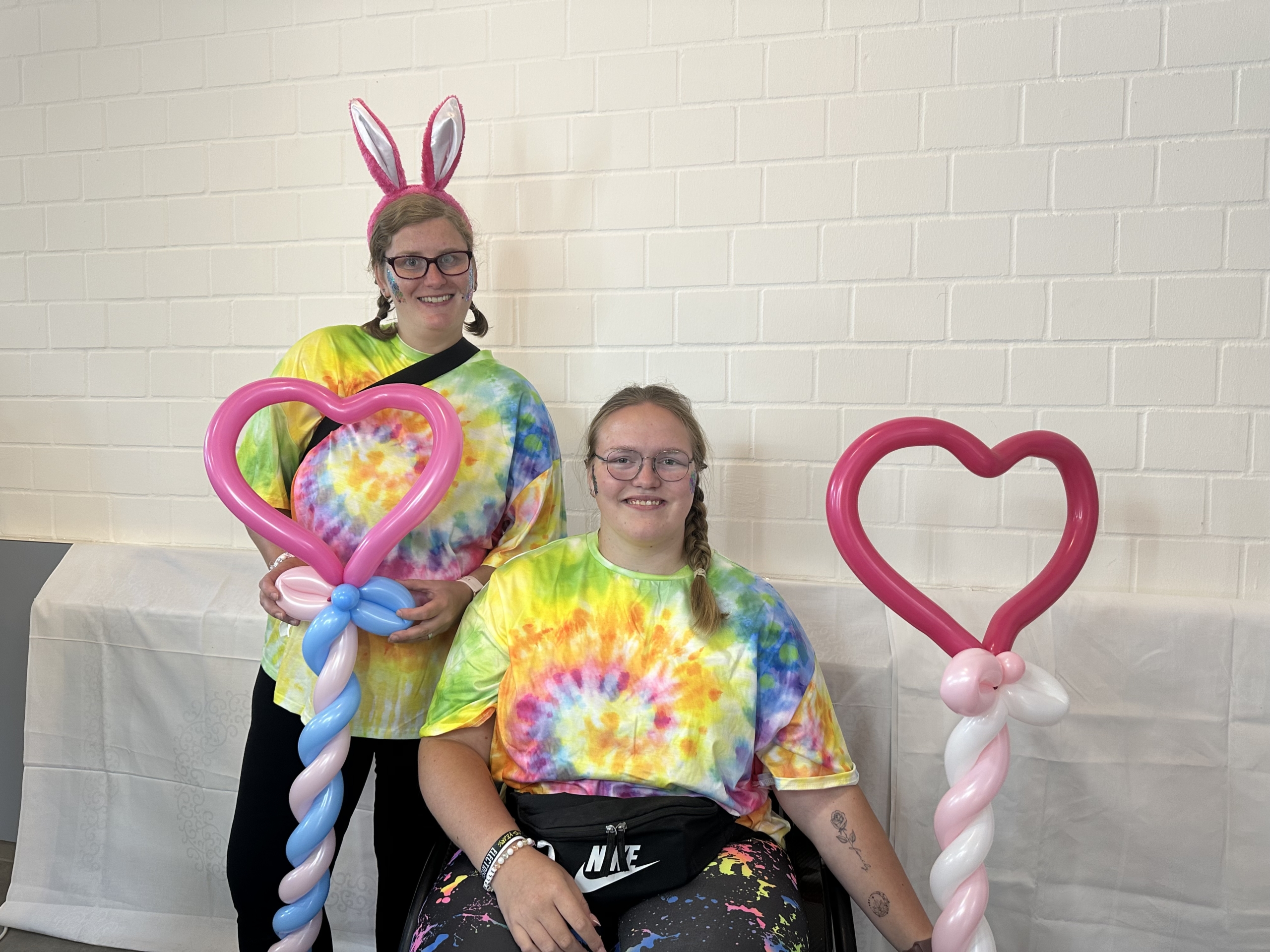 Zwei Personen mit bunten T-Shirts im Batikdesign stehen vor einer hellen Wand. Eine Person trägt Hasenohren und hält einen pinken Herzballon, während die andere einen bunten Stock mit einem großen Herz in der Hand hält.