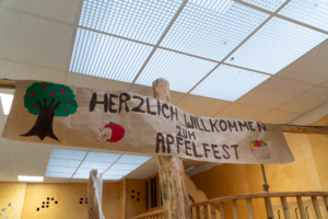 Banner mit der Aufschrift "Herzlich Willkommen zum Apfelfest", dekoriert mit Symbolen und Farben, die die Festlichkeit des Ereignisses unterstreichen.