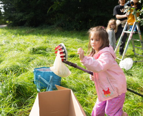 Ein Mädchen in einem pinken Pullover spielt im Freien mit einem Spielzeug, während andere Kinder im Hintergrund aktiv sind. Die Szene findet auf einer grünen Wiese statt, was eine fröhliche und spielerische Atmosphäre vermittelt.