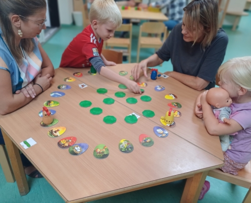 Kinder und Erwachsene spielen gemeinsam an einem Tisch mit bunten Spielsteinen und einem Spielbrett. Die Szene zeigt eine förderliche Lernumgebung, die soziale Interaktion und Teamarbeit unterstützt.