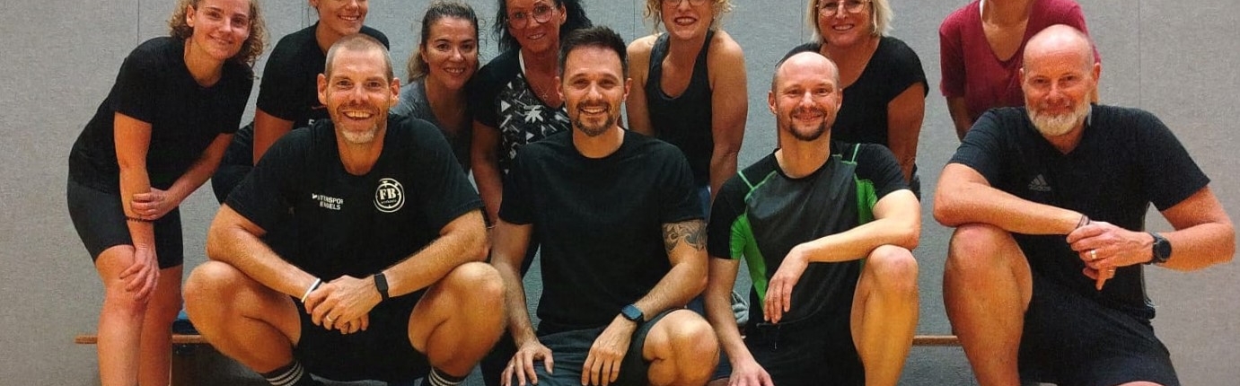 Gruppe von Sportlern in sportlicher Kleidung, die lächelnd auf dem Boden sitzen, in einer Turnhalle.