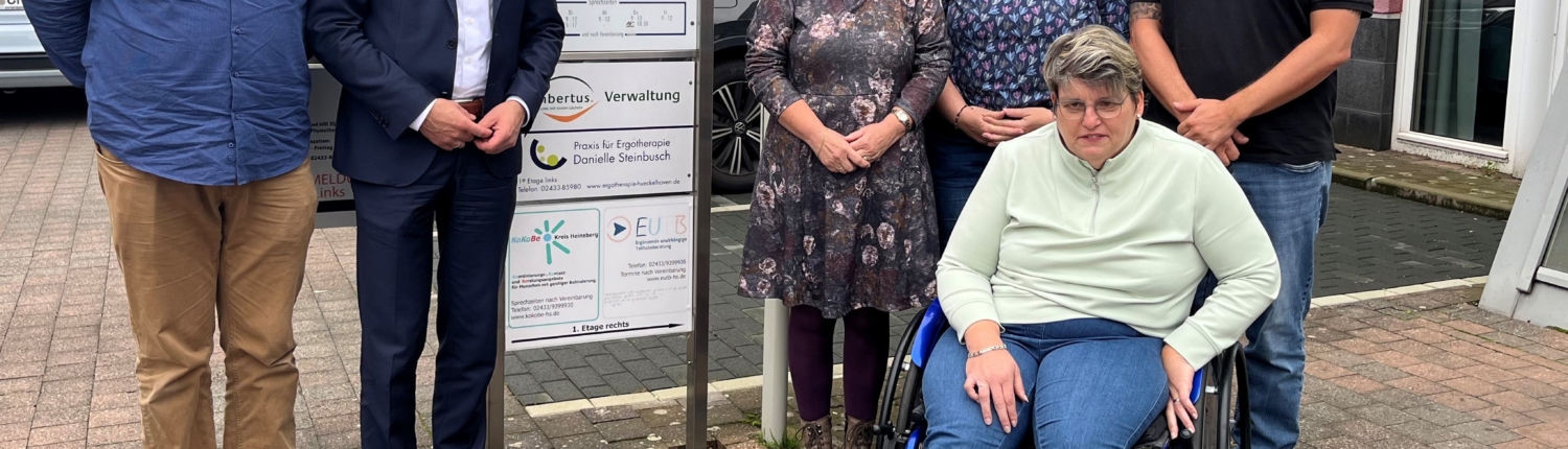 Gruppe von fünf Personen, darunter eine Frau im Rollstuhl, posiert vor einem Informationsschild in einer Stadt. Sie stehen für ein gemeinsames Projekt oder eine Veranstaltung.