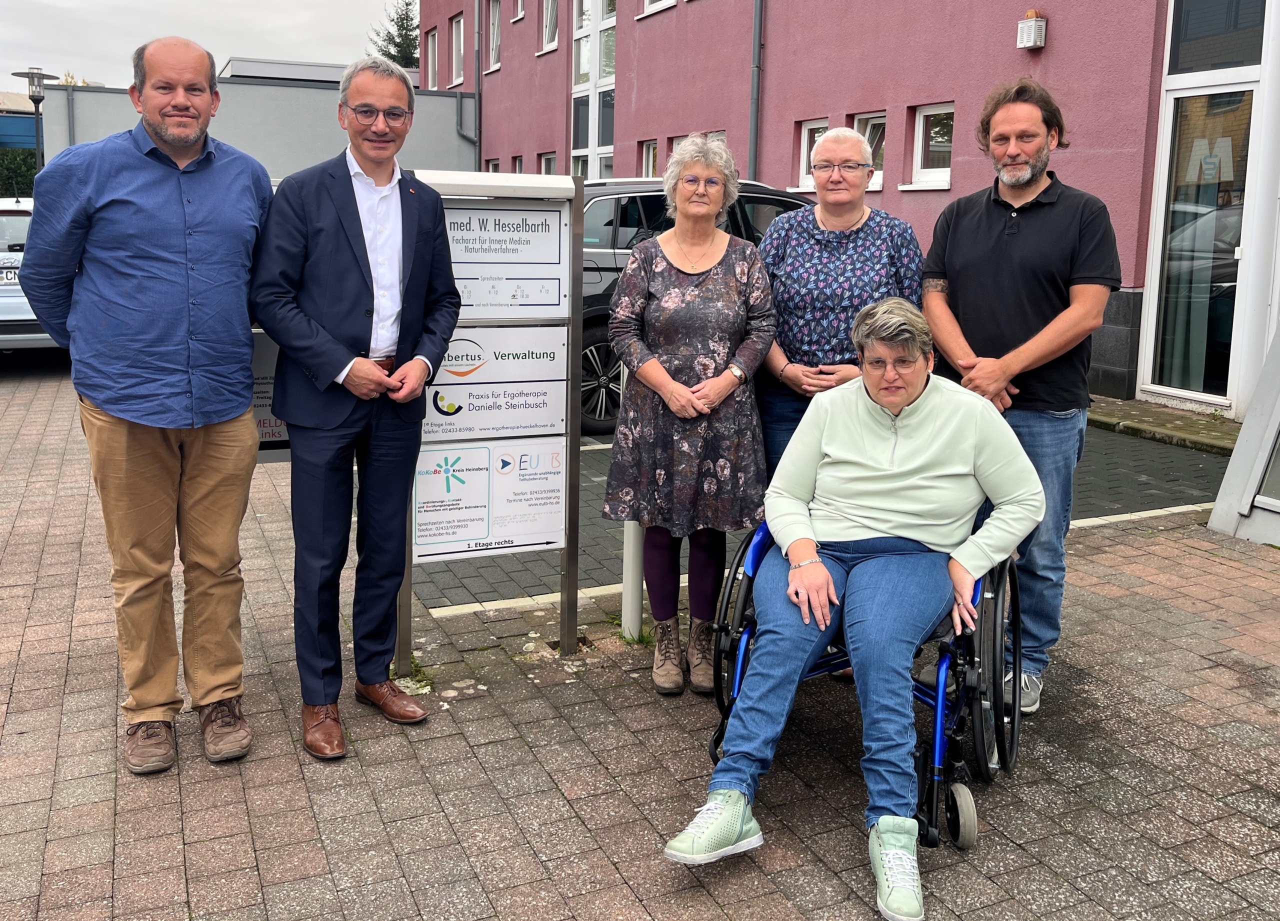 Gruppe von fünf Personen, darunter eine Frau im Rollstuhl, posiert vor einem Informationsschild in einer Stadt. Sie stehen für ein gemeinsames Projekt oder eine Veranstaltung.