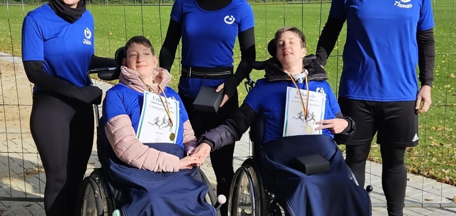 Gruppe von fünf Menschen, darunter zwei Rollstuhlfahrer, die in blauen Sportanzügen im Freien stehen. Sie lächeln und posieren gemeinsam für ein Foto, um ihre Teilnahme an einer Sportveranstaltung für Menschen mit Behinderungen zu zeigen.