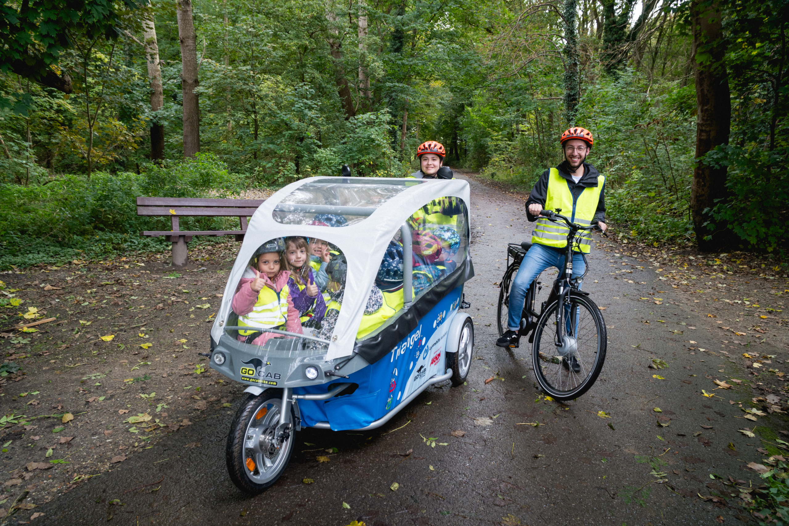 Eine Familie fährt mit einem Dreirad im Wald. Die Kinder sitzen vorne, die Eltern hinten. Alle tragen Helme und leuchtende Westen.