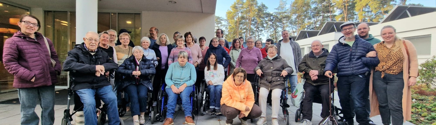 Gruppe von Menschen, die im Freien stehen und zusammen posieren, vor einem Gebäude mit Bäumen im Hintergrund. Die Szene zeigt eine vielfältige Gemeinschaft, die gemeinsam Zeit verbringt.