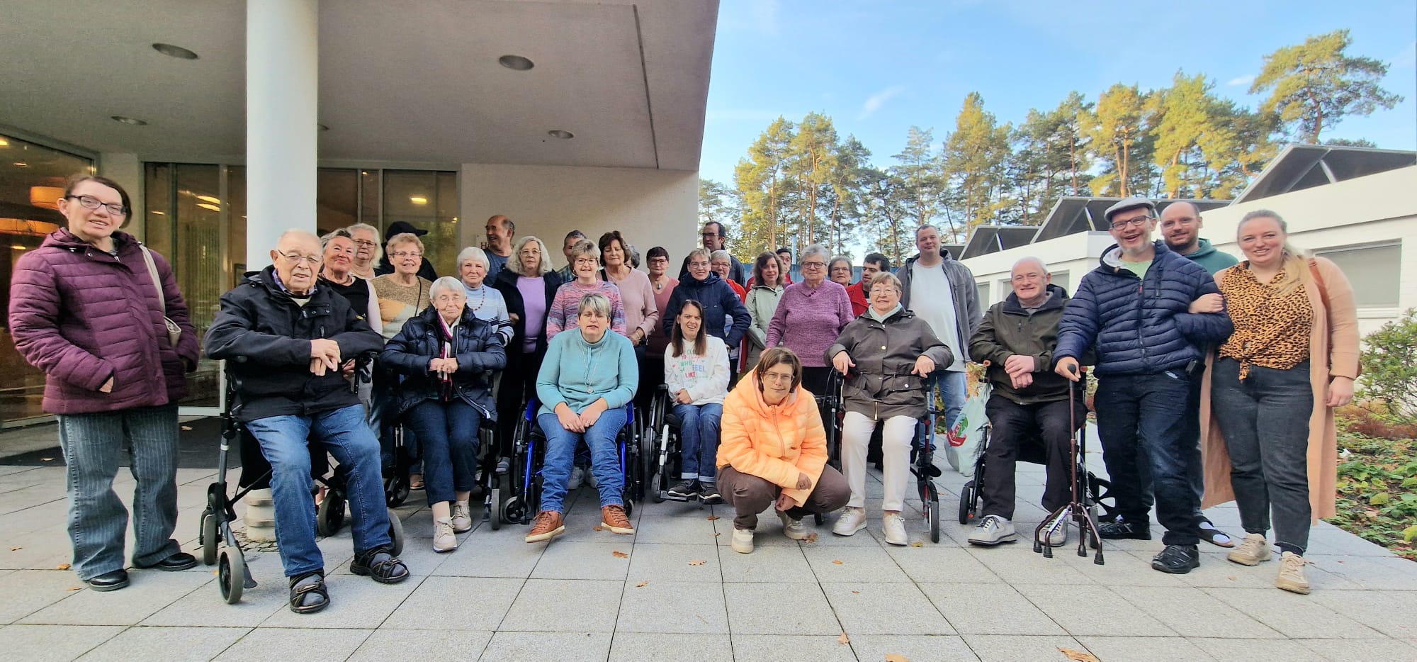 Gruppe von Menschen, die im Freien stehen und zusammen posieren, vor einem Gebäude mit Bäumen im Hintergrund. Die Szene zeigt eine vielfältige Gemeinschaft, die gemeinsam Zeit verbringt.