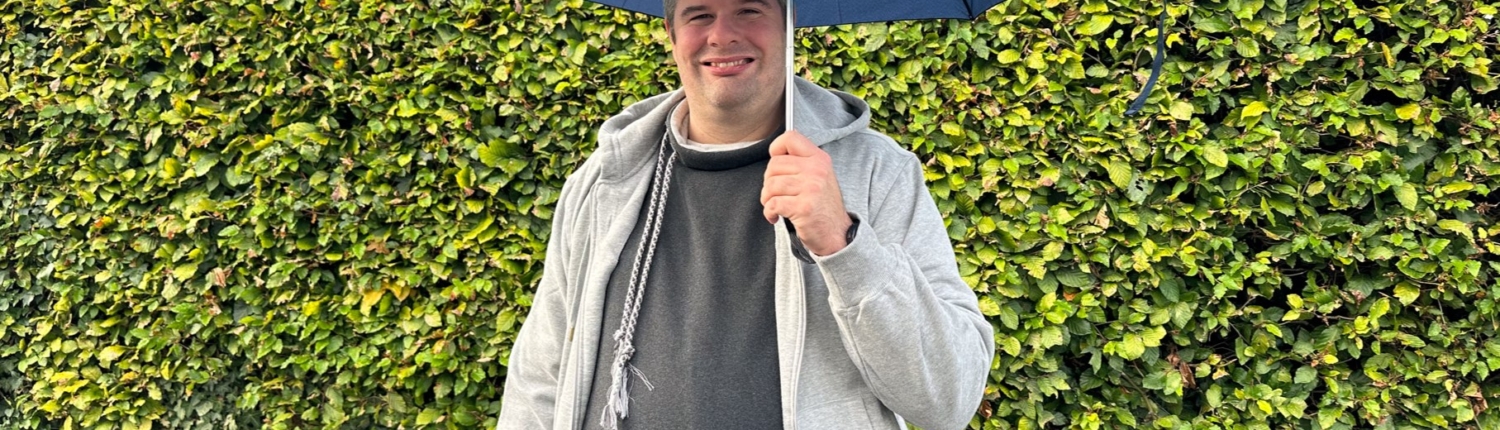 Mann mit blauer Regenschirm steht vor einer grünen Hecke und trägt einen grauen Pullover.