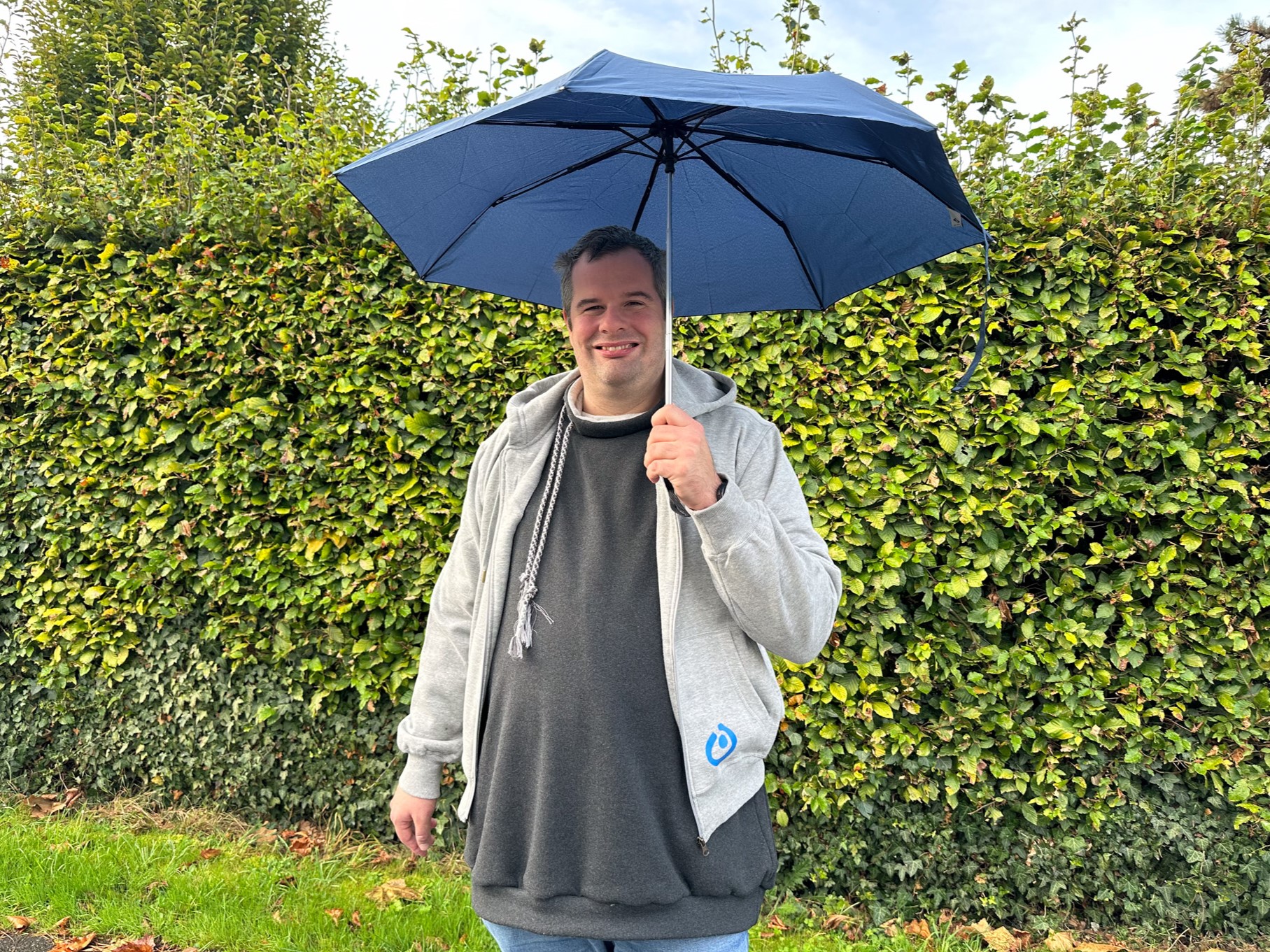 Mann mit blauer Regenschirm steht vor einer grünen Hecke und trägt einen grauen Pullover.