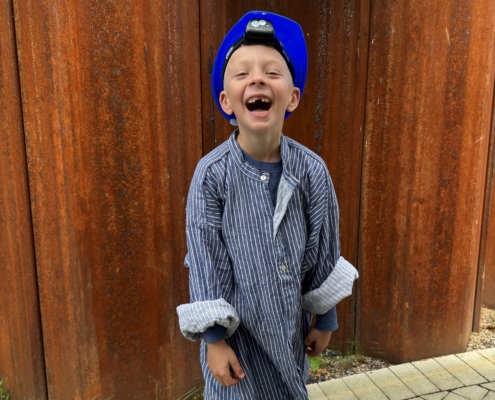 Junge mit blauer Mütze und traditioneller Kleidung, der fröhlich im Freien lacht, vor einer Holzwand stehend.