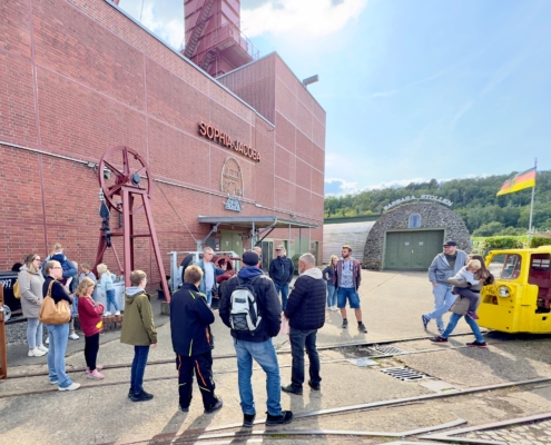 Besuchergruppe vor einer historischen Industrieanlage, umgeben von roten Backsteinmauern und Maschinen, während einer Besichtigung oder Veranstaltung im Freien.