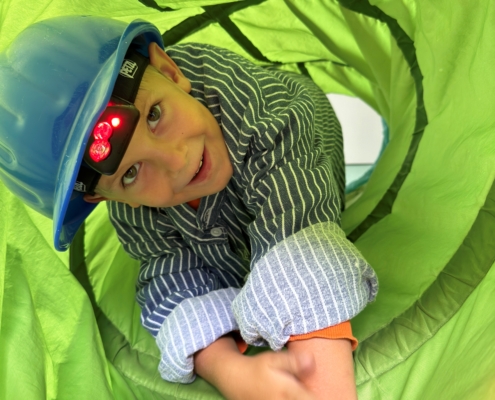 Ein Kind mit einem blauen Helm kriecht durch einen grünen Spieltunnel. Die Umgebung ist hell und freundlich, ideal für Abenteuer im Freien.