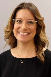 Eine Frau lächelt in die Kamera. Sie trägt eine Brille und hat lockiges Haar.