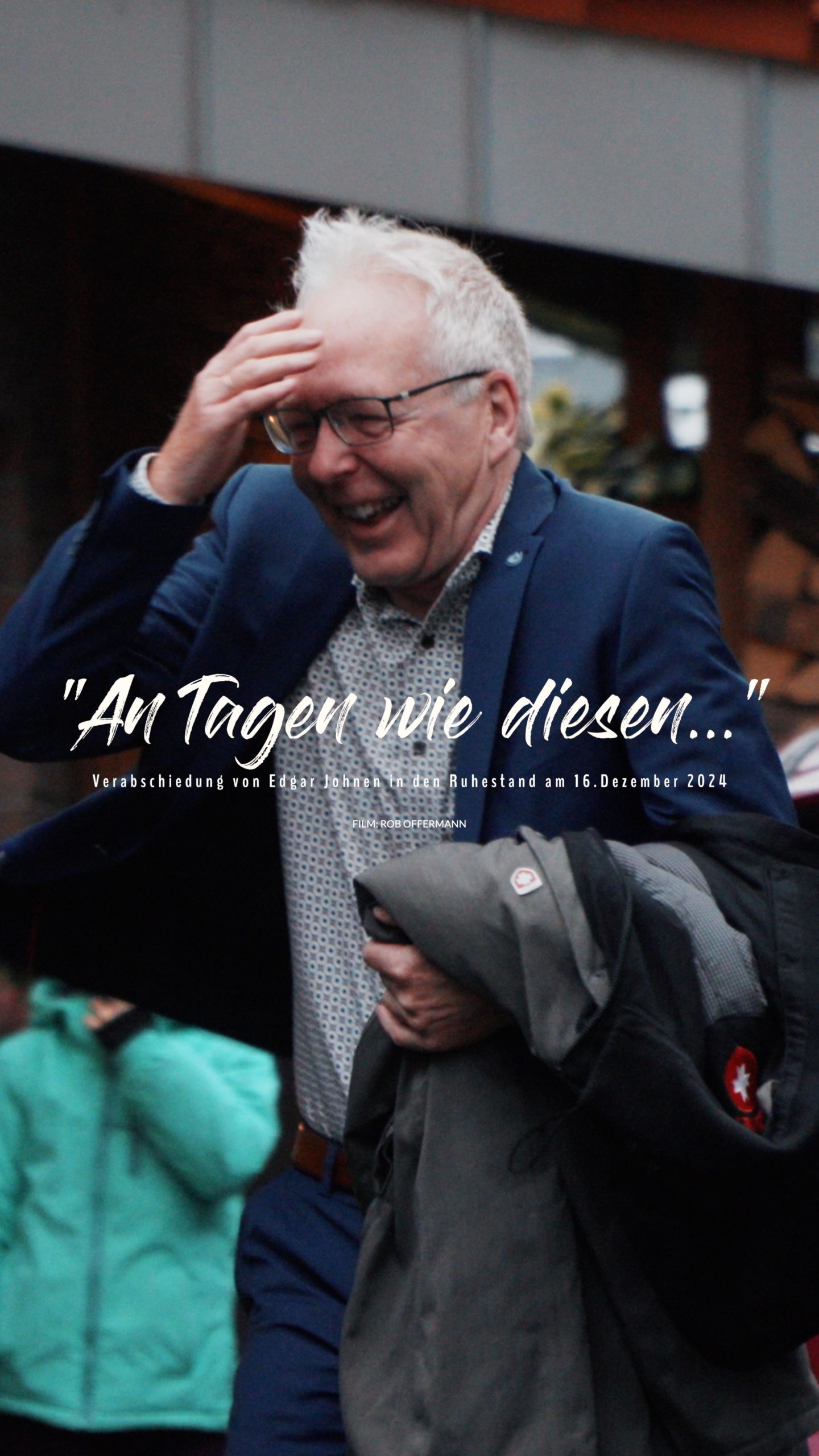 Lächelnder Mann mit Brille, der fröhlich in einer Menschenmenge steht, während er seine Jacke hält. Hintergrund zeigt eine lebhafte Atmosphäre.