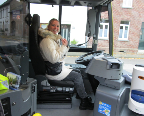 Frau sitzt im Fahrersitz eines Busses und lächelt, während sie auf die Straße schaut. Das Bild zeigt das Innere des Busses mit Steuer und Bedienelementen.