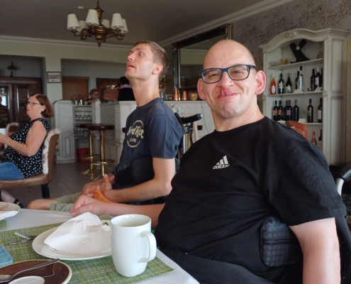 Freundliche Gruppe von Menschen, die an einem Tisch sitzen und gemeinsam eine Mahlzeit genießen. Der Fokus liegt auf einem Mann mit Brille, der lächelt, während andere im Hintergrund entspannt sind.