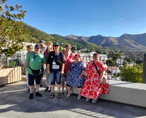 Gruppe von acht Personen in bunten Outfits, posierend auf einer Terrasse mit Blick auf Berge und blauen Himmel.