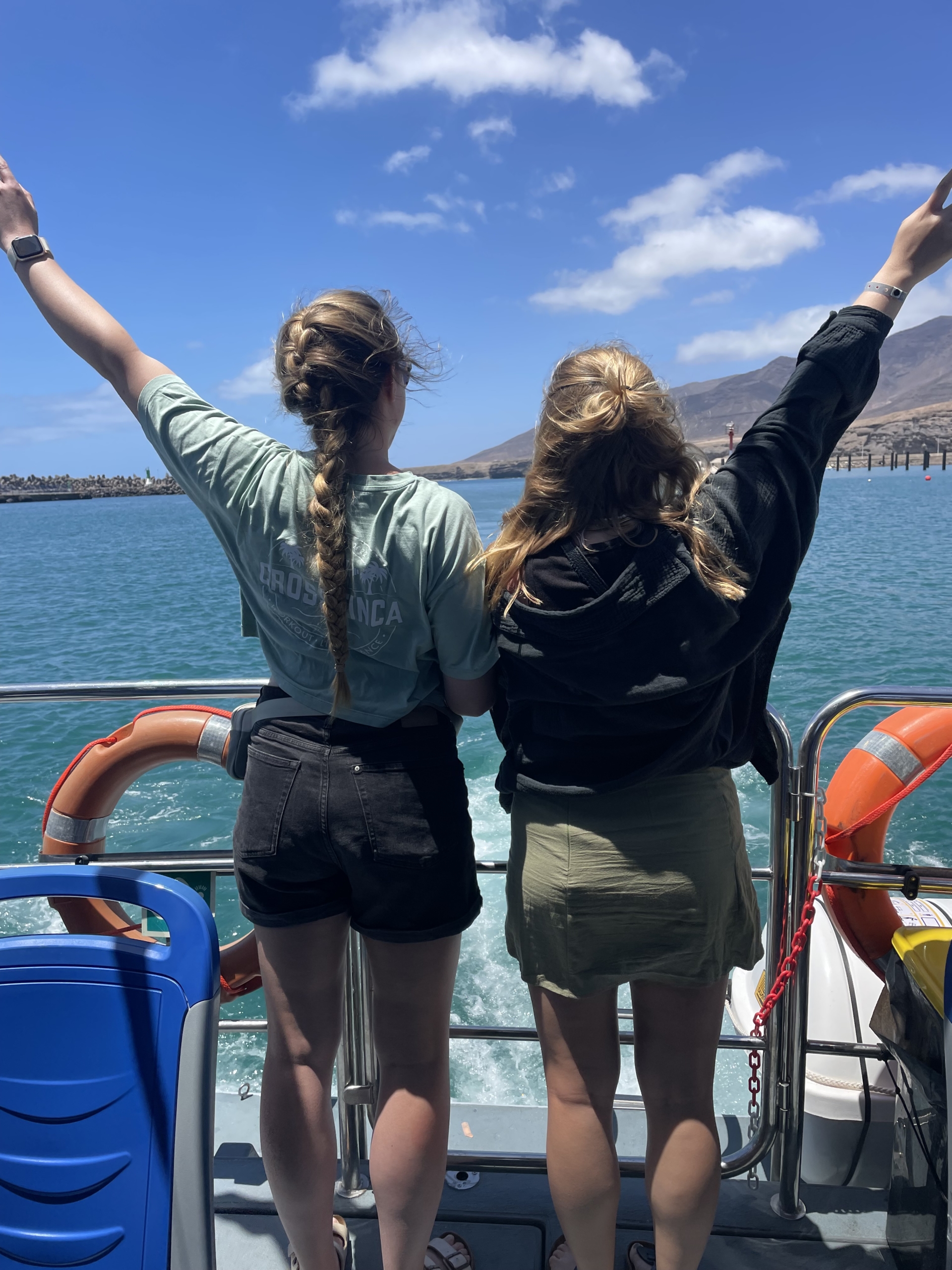Zwei Frauen stehen auf einem Boot und genießen die Aussicht auf das Wasser, während sie ihre Arme in die Luft heben. Im Hintergrund sind blauer Himmel und Berge sichtbar.