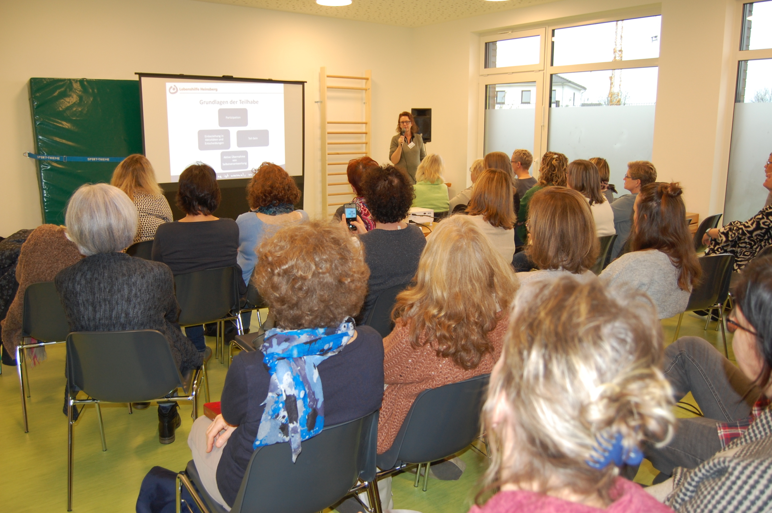 Eine Gruppe von Menschen sitzt in einem Seminarraum und hört einem Vortrag zu, während eine Präsentation an die Wand projiziert wird.