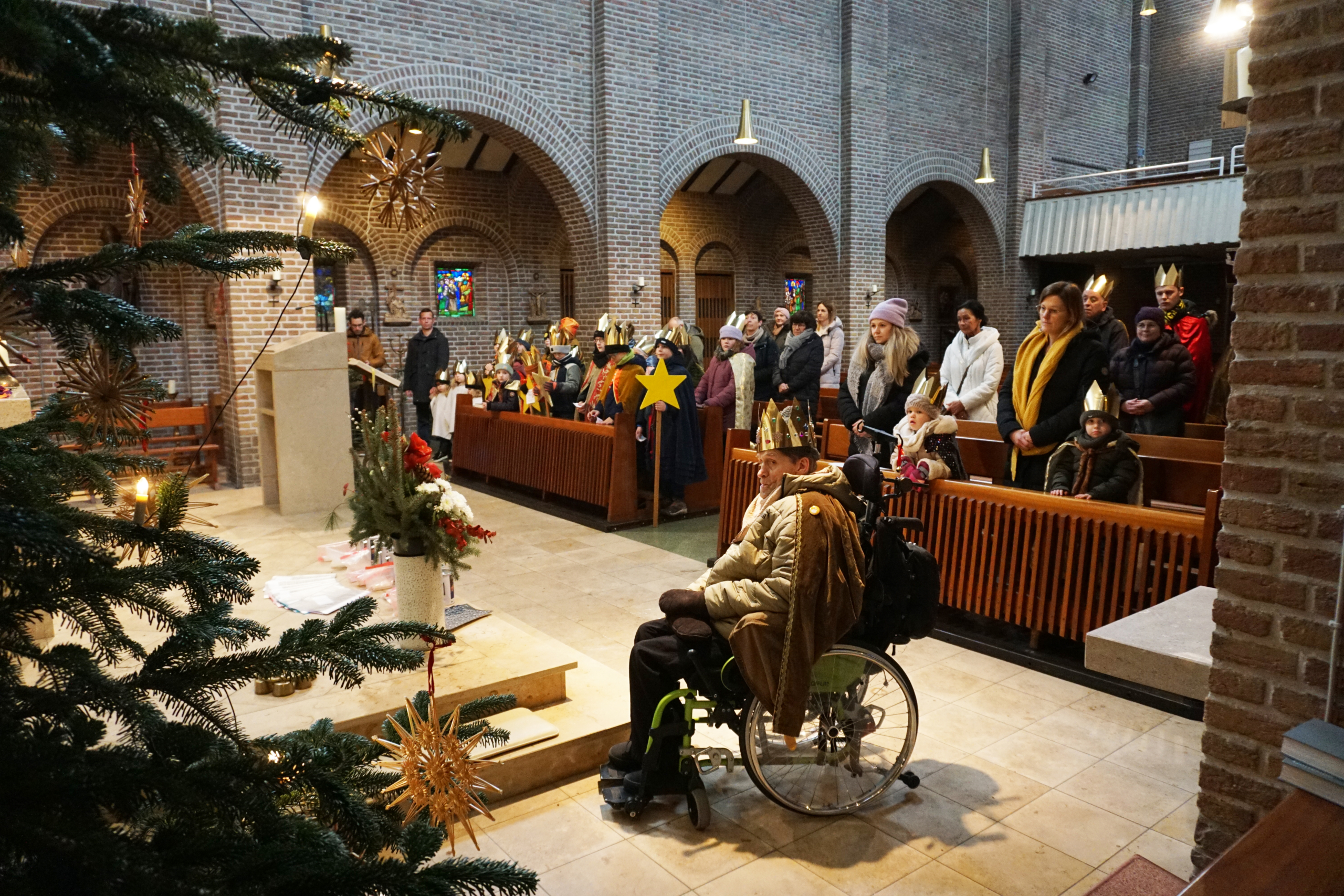 Weihnachtsfeier in einer Kirche. Menschen sitzen auf Bänken. Ein Mann sitzt im Rollstuhl. Es gibt einen Weihnachtsbaum.