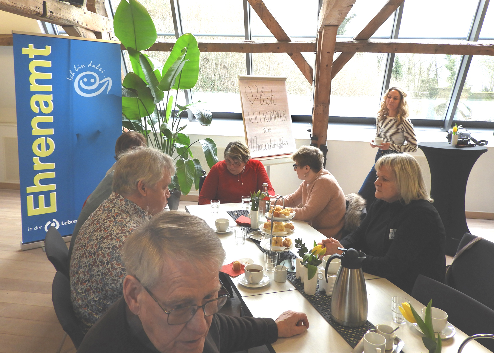 Eine Gruppe von Menschen bei einem Treffen in einem hellen Raum mit großen Fenstern. Sie diskutieren und arbeiten an einem Tisch, umgeben von Pflanzen und Arbeitsmaterialien.