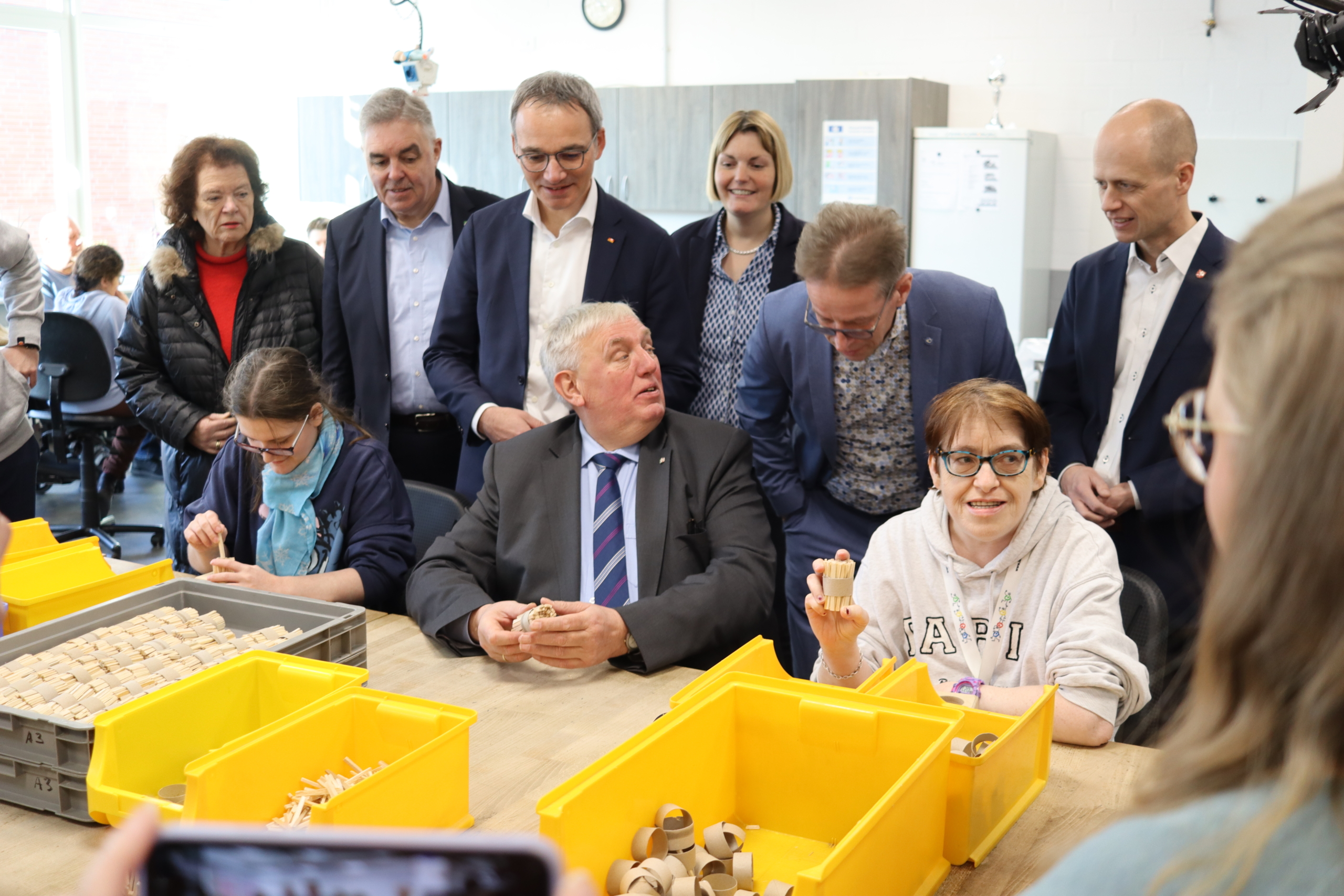 Männer und Frauen stehen in einem Raum. Sie schauen auf Holzstücke in gelben Kisten.