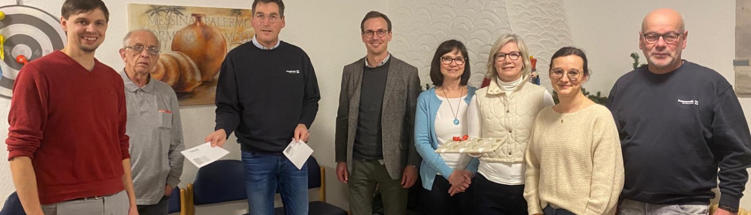 Eine Gruppe von Menschen steht in einem Raum. Einige tragen Jacken oder Pullover. Im Hintergrund sind Regale mit Lebensmitteln zu sehen.