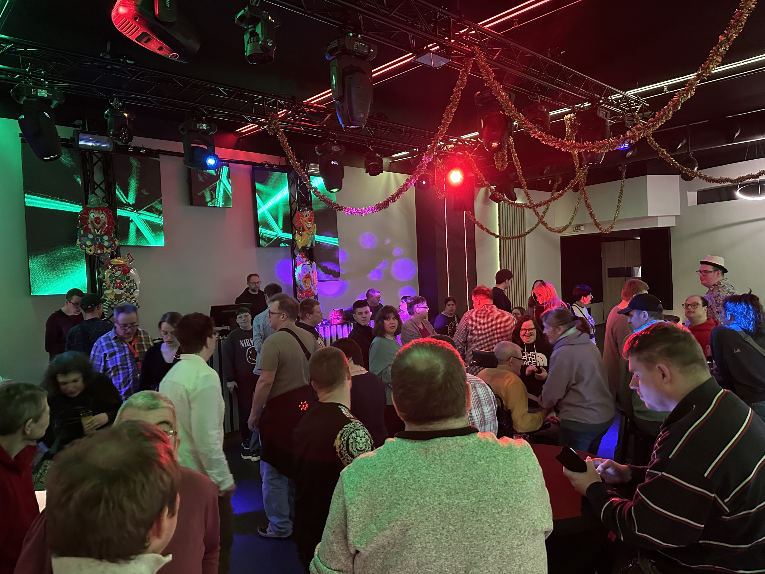 Viele Menschen tanzen in einem Raum. Es gibt bunte Lichter und Musik.